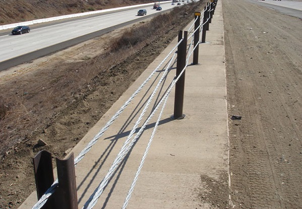 View of a multi-late freeway from the center median. The median divider is made of large steel posts with multiple steel wires woven between them.