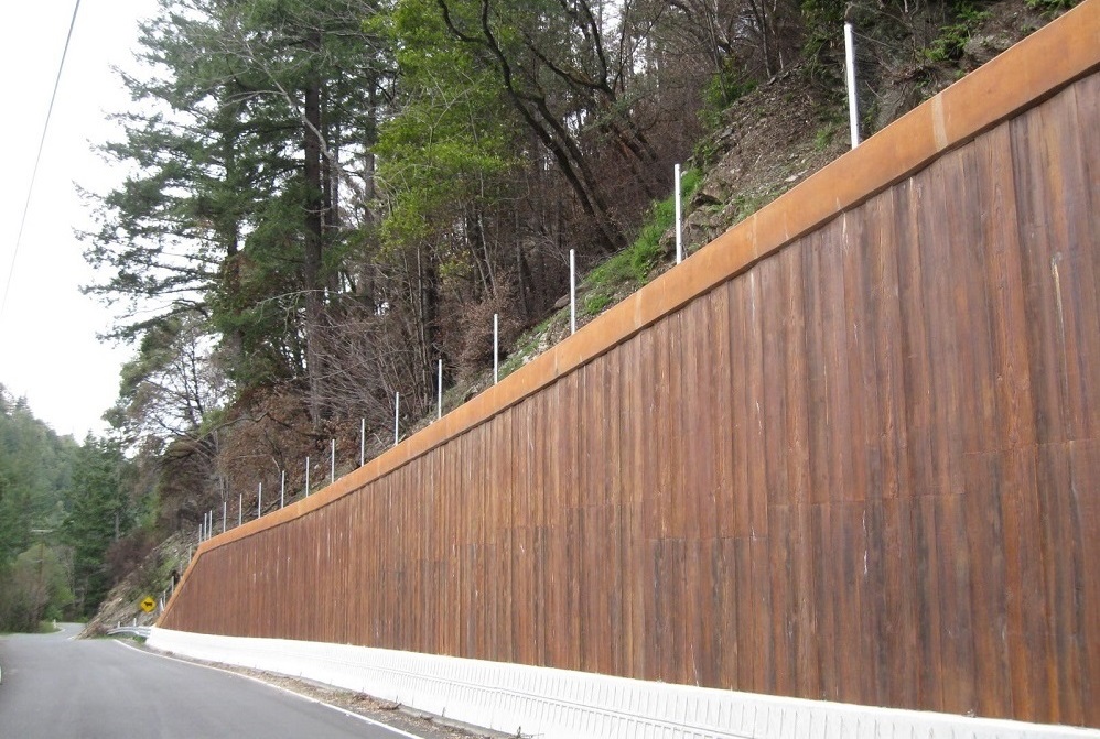 A view of a plank wooden wall with a hillside, trees and shrubs above and behind the wall.