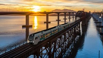 Color image at sunset of a train on the Benicia Bridge, surrounded by water below. The white train has blue graphics that indicate it is a "Zero Emission" passenger train and Caltrans logos are visible on the front and side.
