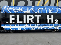 A color image of a hydrogen-fuel-powered passenger train. The train wrap has a blue and white pattern and the words "FLIRT H2" which stands for Fast Light Intercity and Regional Train. Project partner logos for the California State Transportation Agency (CalSTA), the San Bernardino County Transportation Authority (SBCTA), Metrolink and Caltrans are visible.
