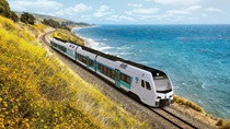 Color image of a train on the California coast, with a yellow-flower-covered hillside above to its left and the blue Pacific Ocean to its right below. The white train has blue graphics that indicate it is a "Zero Emission" passenger train and Caltrans logos are visible on the front and side.