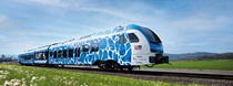 A color image of a hydrogen-fuel-powered passenger train traveling through a field of green grass and white flowers. The train wrap has a blue and white pattern that also reads, "FLIRT H2." On the front right side of the train below an American flag graphic, project partner logos for the California State Transportation Agency (CalSTA), the San Bernardino County Transportation Authority (SBCTA), Metrolink and Caltrans are visible. On the front, a logo for train builder Stadler Rail, Inc. is visible.