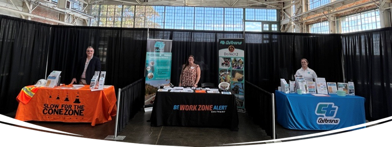 Three Caltrans staff conduct outreach at a small business event, each standing behind a table with various pamphlets and fliers. They're set up in a large room with floor to ceiling windows. Each table is covered by a tablecloth. One reads Slow for the Cone Zone, another reads Be Work Zone Alert and the third reads Caltrans. 