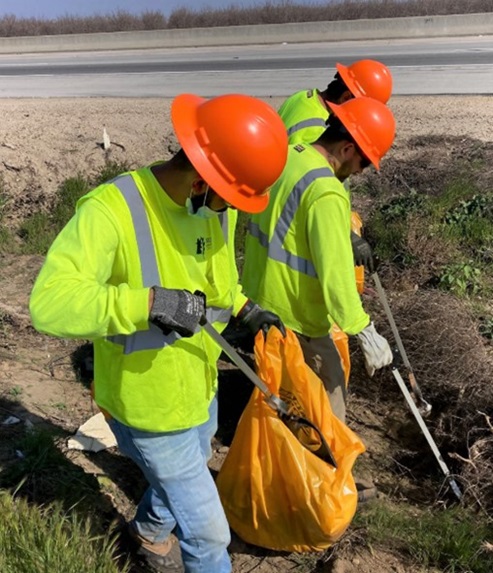 Three workers wearing gloves, orange hardhats, and fluorescent yellow shirts and vests use grabber tools to pluck roadside trash off the ground for removal in orange garbage bags, as they work to beautify the highway,