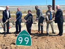 Six state and local officials pose for a photo while digging dirt with ceremonial shovels behind a green and white "California 99" State Highway Route sign.