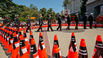Photo of the 2022 Caltrans Workers Memorial Ceremony. Orange cones with black name bands representing the 189 fallen state highway workers who died in the line of duty since 1921 are arranged in a diamond shape like a caution sign. Inside the diamond, uniformed Caltrans Honor Guards place a ceremonial black cone in front of a floral wreath during the memorial. 