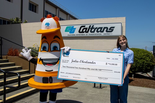 Cone-shaped Caltrans mascot Safety Sam holds one side of a large novelty check displaying the name of Julia Oberlander and the amount of $2,250 scholarship recipient Julia holding the other side. 