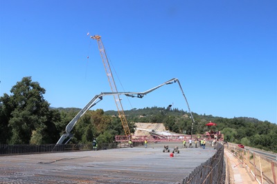 View from the bridge deck of Calpella Two Bridge Replacements Project. A cement crane is in the background with contractor crews working in the foreground. 