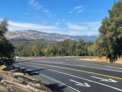 Intersection of two roads, Road 144 and State Route 20 in Mendocino County. Trees line the road on all sides. Bright white and yellow lane markers outline the two-lane highway and offramp and onramp to Road 144.