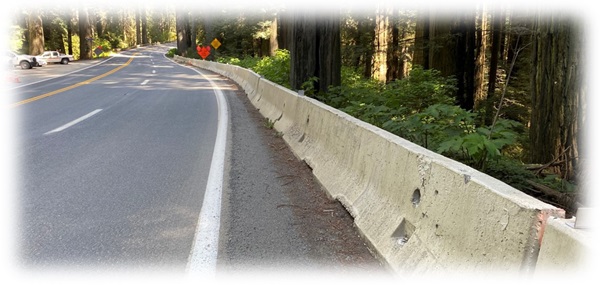 Three lanes of U.S. 101 in Del Norte County extend into the background. The road is rimmed with concrete barriers. Pickups are visible in an off-street dirt parking lot, and traffic control signs are visible along the road in the distance. 