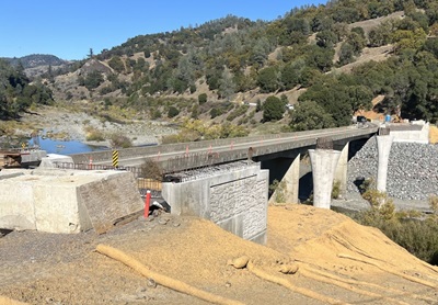 Construction of Eel River Bridge Replacement in Mendocino County. New columns are visible on one side of the existing bridge and new abutments are in place on either side of the river. A gently sloping mountainside frames the roadway and the river extends beyond the bridge. 