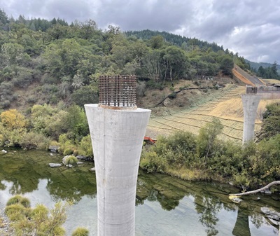 Construction of Eel River Bridge Replacement in Mendocino County. New columns are visible on one side of the existing bridge and new abutments are in place on either side of the river. A gently sloping mountainside frames the roadway and the river extends beyond the columns. 