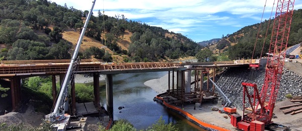 Construction of Eel River Bridge Replacement in Mendocino County. New columns are visible on one side of the existing bridge and new abutments are in place on either side of the river. A gently sloping mountainside frames the roadway and the river extends beyond the bridge.