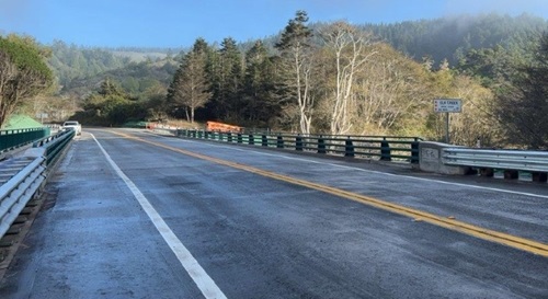 The elk creek bridge deck is shining from recent rain though the sun shines in the background, spotlighting a small forest adjacent to the roadway. 