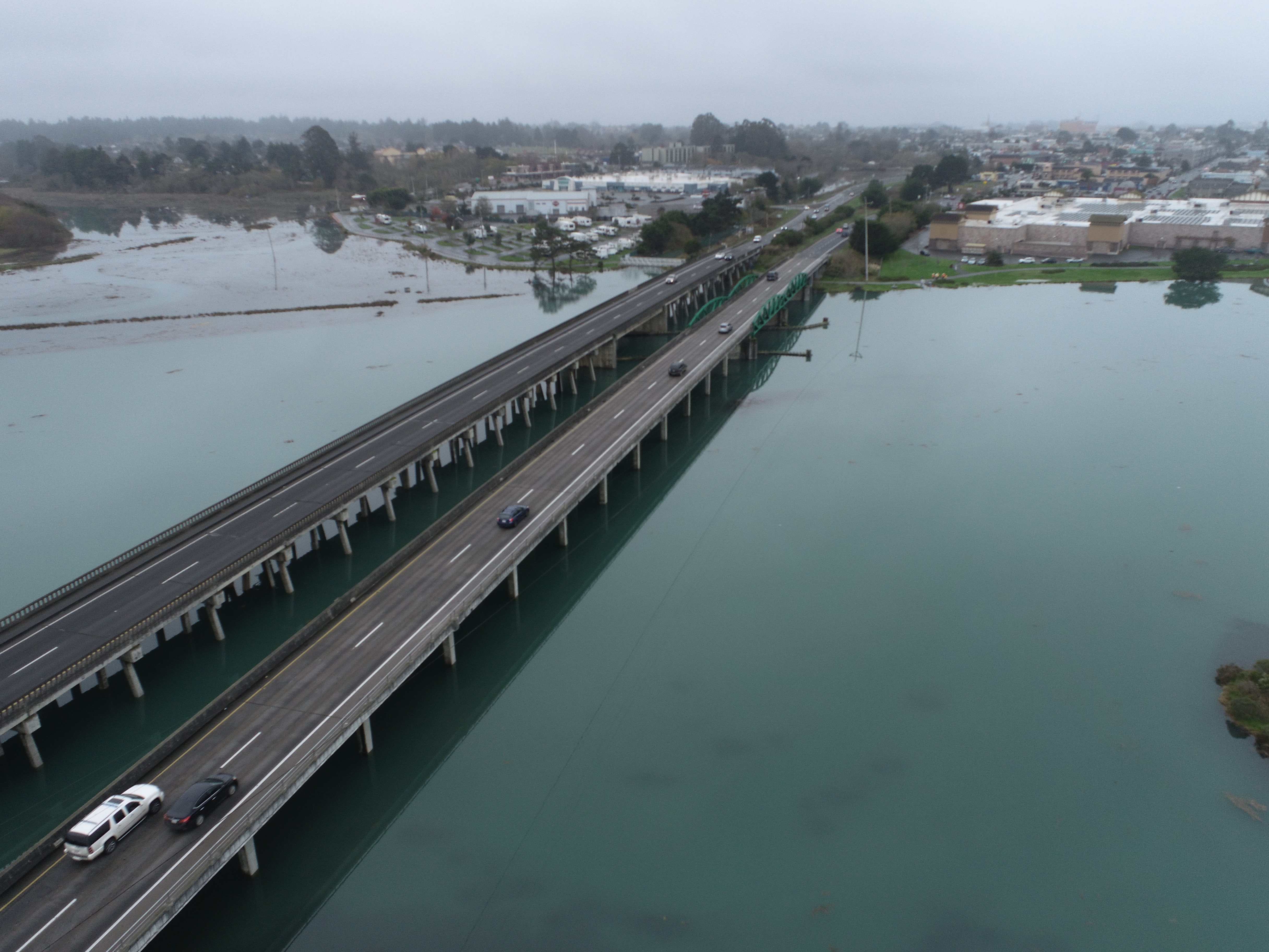 Aerial view of the Eureka Slough Bridge looking southeast. Vehicles travel on the bridge deck. Concrete pillars extend from under the bridge deck into the green water of the Eureka Slough. The northern edge of Eureka is visible in the background.