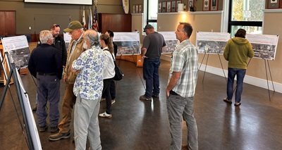 A group of residents assemble in a large conference room with displays on easels. Caltrans staff talk with a few attendees as others view the displays with renderings of the Fort Bragg ADA project. 