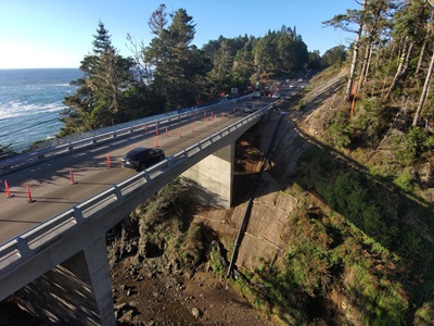 Aerial view of the new jack peters creek bridge. The new pavement and columns are bright grey and the creekbed below is dry. A single car travels away from the camera, navigating through cones  indicating one-way traffic control. 