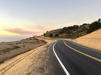 Rebuilt and repaved two-lane section of Route 1 in Mendocino County. The ocean is visible in the background under an orange and blue sky. 