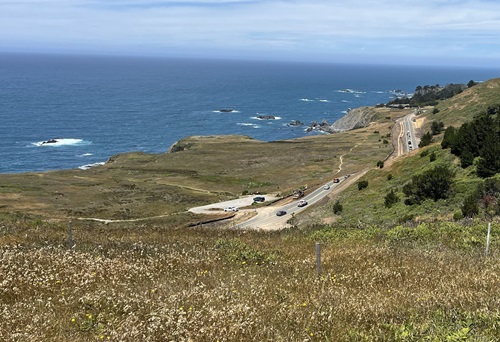 A sunny day along Route 1 on the Mendocino Coast. A hill slopes down to a narrow roadway. Heavy equipment is positioned in multiple positions while vehicles traverse the highway.