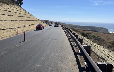 Repaved roadway with two-way traffic. Erosion control barriers line the hillsides parallel to the road, and guardrail separates the lanes from long flat land that slopes into the ocean. The pacific glints in the sunlight under a blue sky.