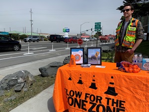 Caltrans staff in a reflective safety vest stands behind a table at the intersection of Broadway and Wabash in Eureka California. Safety materials are arranged on the table alongside educational pamphlets describing the Broadway Pop-Up Demonstrations. 