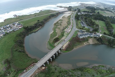 Aerial view of the Pudding Creek Bridge on Route 1 near Fort Bragg. A two-lane bridge spans a creek and watershed with the ocean outlet visible in the background. Houses line the creek on the east side and between the creek and ocean on the westside. 