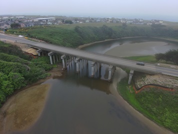 Aerial view of Pudding Creek Bridge looking west on a foggy day. The current bridge's seven column supports are visible in the low water level of the creek. Erosion control barriers snake along the hillside under the bridge. A single southbound car populates the bridge. 