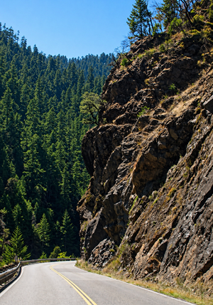 A rocky outcropping hangs above the U.S. 199 roadway. The two-lane road hugs the cliff and the guardrail with no shoulder on either side. 