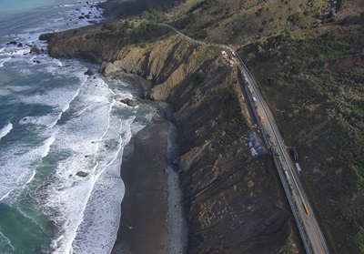 aerial view of State Route 1 in Mendocino County. The road is flanked by a vegetation covered hillside and bordered by white capped waves.