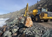 Heavy equipment moves large boulders to fortify the slope under Route 1 in Mendocino County. The ocean and rocky hillside are visible in the background. 