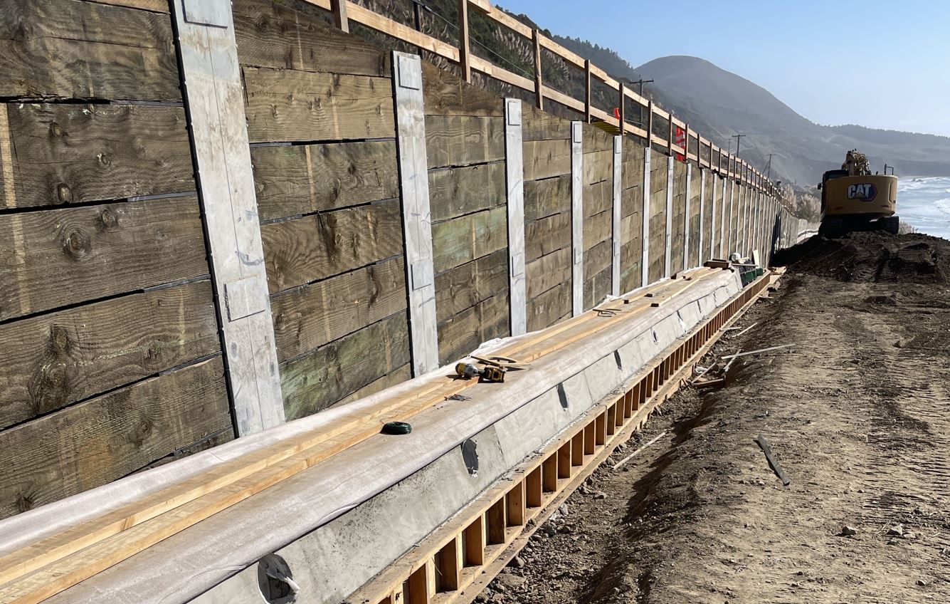 Retaining wall on Route 1 in Mendocino County. Wood is laid parallel to the ground; slid into concrete forms. An excavator crests a pile of dirt with the blue ocean visible under behind under a hazy sky.