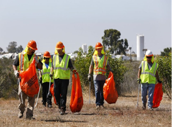 Group of people walking, wearing safety helmets and reflective vests, holding orange trash bags. For more information, call (619) 688-6670 or email CT.Public.Information.D11@dot.ca.gov
