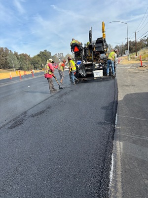 Crews laying asphalt along State Route 49 as part of a safety barrier project.