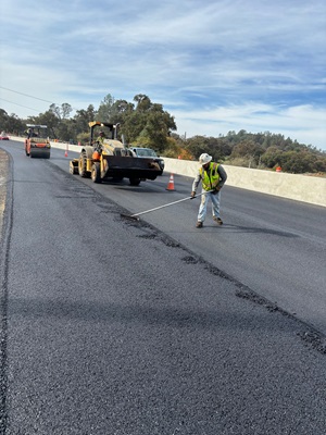 Crews laying asphalt along State Route 49 as part of a safety barrier project.