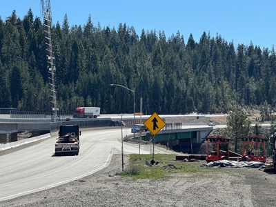 Construction zone at the Yuba Pass Separation Overhead Bridge Project along I-80 in Nevada County, showing a gravel staging yard leading toward an elevated highway. A yellow merge‑lane sign is visible in the center. Trucks travel on both the lower and the upper highway structure. Construction equipment, materials, and a tall crane are positioned near the bridge. A forested hillside forms the background under clear blue skies.