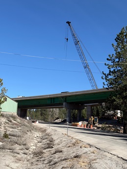A highway overpass under construction with green steel beams supported by concrete columns. A tall crane stands beside the structure, and construction equipment and orange traffic cones are visible on the ground below. The scene includes dry roadside vegetation, a small green building on the left, and tall trees in the background under a clear blue sky.