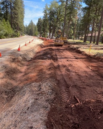 A road construction site beside a two‑lane rural road lined with orange cones. A section of earth next to the roadway has been excavated, showing reddish dirt and tire tracks. A piece of heavy machinery is parked further ahead, and tall pine trees surround the area under a sunny sky.