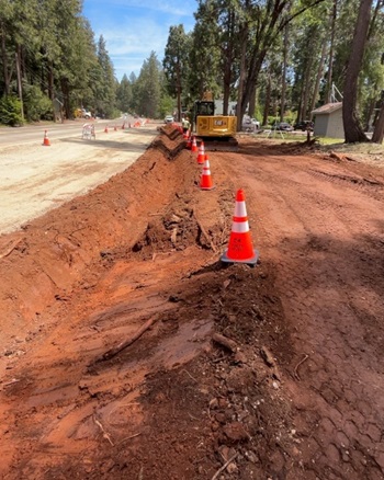 A roadside construction area where a long trench of reddish soil has been excavated beside a two‑lane rural road. Orange traffic cones line the edge of the trench, guiding vehicles away from the work zone. A yellow CAT excavator sits further up the cleared dirt area near tall pine trees, with houses partially visible in the background under a sunny sky.