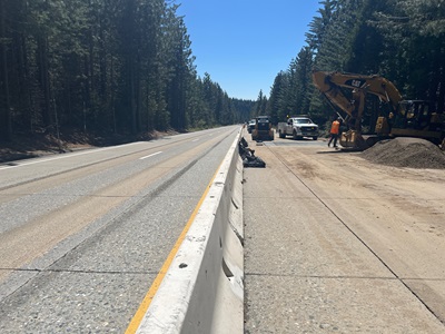 A highway work zone in a forested area. A concrete barrier separates the open traffic lanes on the left from the construction area on the right. Several pieces of heavy equipment, including a large CAT excavator and compactors, are parked on the work side. A worker in an orange safety vest stands beside a pile of gravel near utility trucks under bright, sunny conditions.