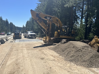 A highway construction zone bordered by tall pine trees. A large yellow CAT excavator sits on the right side near a mound of gravel, with its bucket raised. A work truck and compacting equipment are parked ahead on the dirt work area. On the left, traffic travels past a concrete barrier separating vehicles from the construction site under bright sunlight.