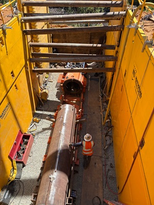 A construction worker wearing a reflective safety vest and hard hat stands inside a deep, narrow trench supported by large yellow shoring walls. Metal cross‑braces span the trench overhead, and a long cylindrical steel pipe or casing lies centered on the gravel-covered trench floor. Tools, cables, and equipment are scattered around the work area, and sunlight illuminates parts of the yellow shoring walls and the worker below.