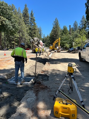 Road construction crews work along a forested two‑lane highway under a clear blue sky. Workers in high‑visibility safety gear stand near a trench filled with fresh concrete while a cement mixer truck pours material into the excavation. An excavator operates farther up the work zone, and construction cones line the area. Surveying equipment, including a yellow laser level on a tripod and a carrying case, is set up near the foreground. Tall pine trees surround the site.