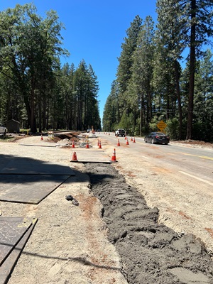 A two‑lane rural road running through a forested area is partially dug up for construction work. Orange traffic cones line both sides of the work zone, and a long trench filled with fresh concrete or fill material runs along the right side of the construction area. Metal plates cover parts of the ground. Several vehicles, including a white truck and a dark car, travel through the single open lane. Tall pine trees border both sides of the road under a clear blue sky.