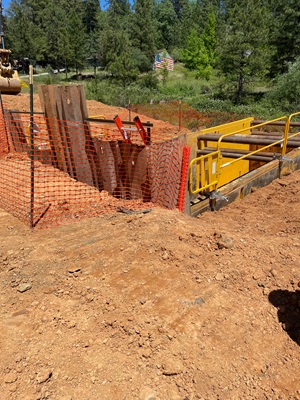 A construction site with reddish dirt and an excavated trench supported by large yellow shoring panels. Orange safety fencing surrounds the area, and steel beams and equipment are positioned inside the excavation. Dense green trees and vegetation fill the background under a clear blue sky.