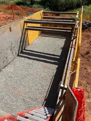 A deep, rectangular trench supported by yellow and gray steel shoring panels is filled with compacted gravel. Several horizontal steel cross‑braces span the width of the trench. A red ladder leads down into the excavation, and orange safety fencing surrounds the work area. Sunlight casts shadows across the gravel surface, with vegetation visible in the background.