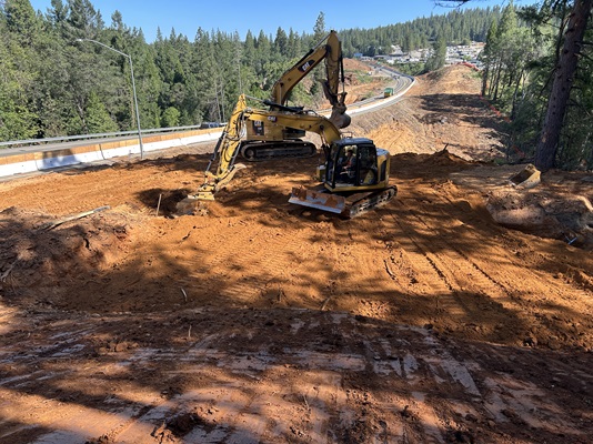 A construction excavator works on a large dirt embankment beside a highway in a forested area. Freshly graded reddish‑brown soil fills the foreground, with the machine smoothing and shaping the terrain. Guardrails and traffic lanes curve through the background, while dense pine trees cover the surrounding hills under a clear blue sky.