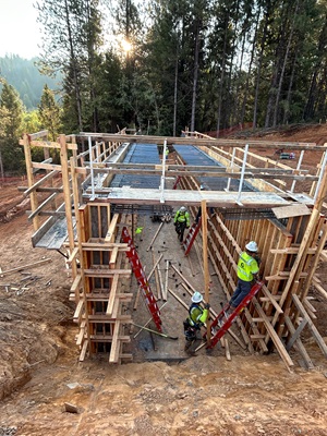 A group of construction workers wearing safety gear assemble tall wooden formwork for a concrete structure in a forested area. The formwork creates a narrow, rectangular passage with red ladders positioned on both sides. Rebar is arranged across the top section, and scattered lumber and tools lie on the ground. Sunlight filters through the surrounding pine trees in the background.