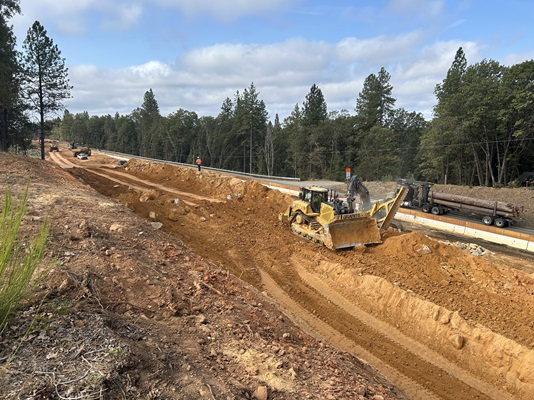 A large yellow bulldozer pushes and levels reddish-brown soil in a wide excavation area along a roadway. Construction vehicles and a worker in an orange vest stand nearby. A truck carrying long logs drives past on the adjacent paved road. The scene is surrounded by tall pine trees under a partly cloudy sky.