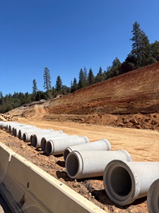 A row of large concrete drainage pipes lies on the ground beside a dirt construction roadway. The area is surrounded by tall pine trees, and a steep, freshly excavated embankment rises to the right. The sky is clear and bright blue, highlighting the open outdoor work site.