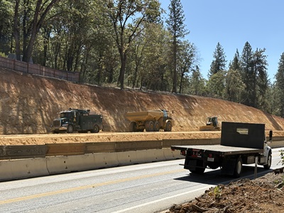 A highway construction site with several large work vehicles operating along a freshly excavated dirt slope. A green service truck, a yellow articulated dump truck, and a small compactor sit on the dirt work area above concrete barriers. A flatbed truck drives along the paved roadway in the foreground. Tall trees line the background under a clear blue sky.